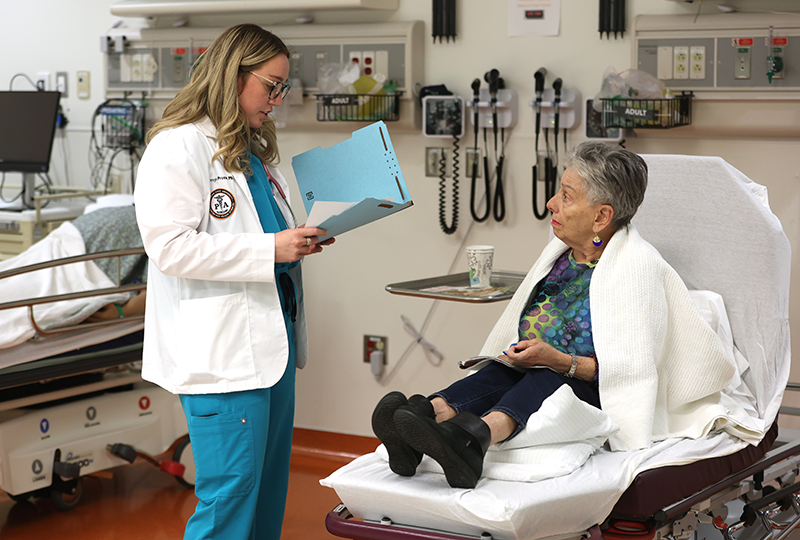 Physician Assistant student holding a folder speaking with a patient sitting on an examination table in the simulation center.
