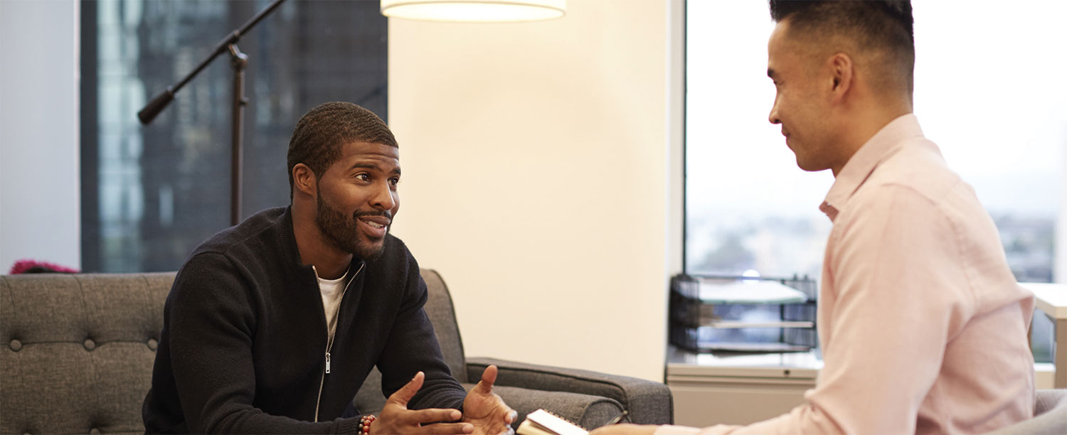 A man sitting on a couch speaking with a man holding a notebook.